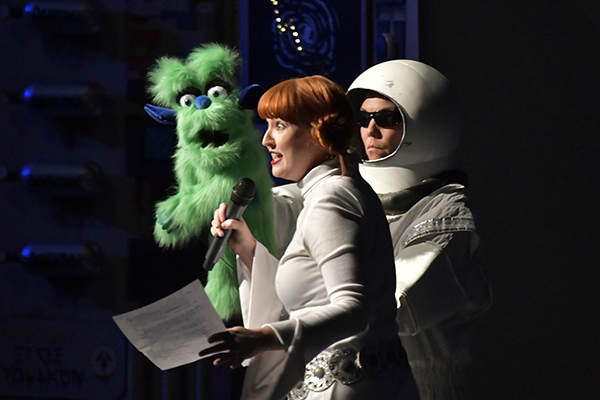 A woman and man dressed in space costumes with a green alien hand puppet