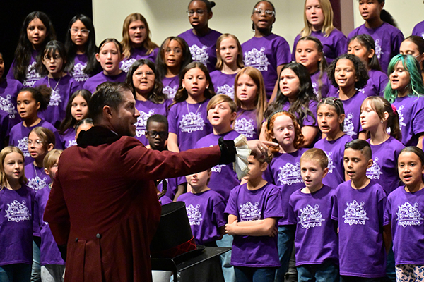 A man directs a group of elementary choir students in purple shirts