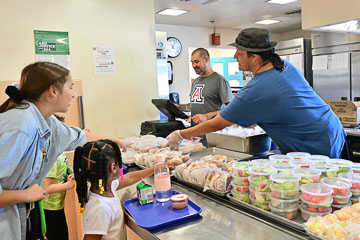 A young student checks out of the cafeteria line with a platter of food and is helped by several staff.