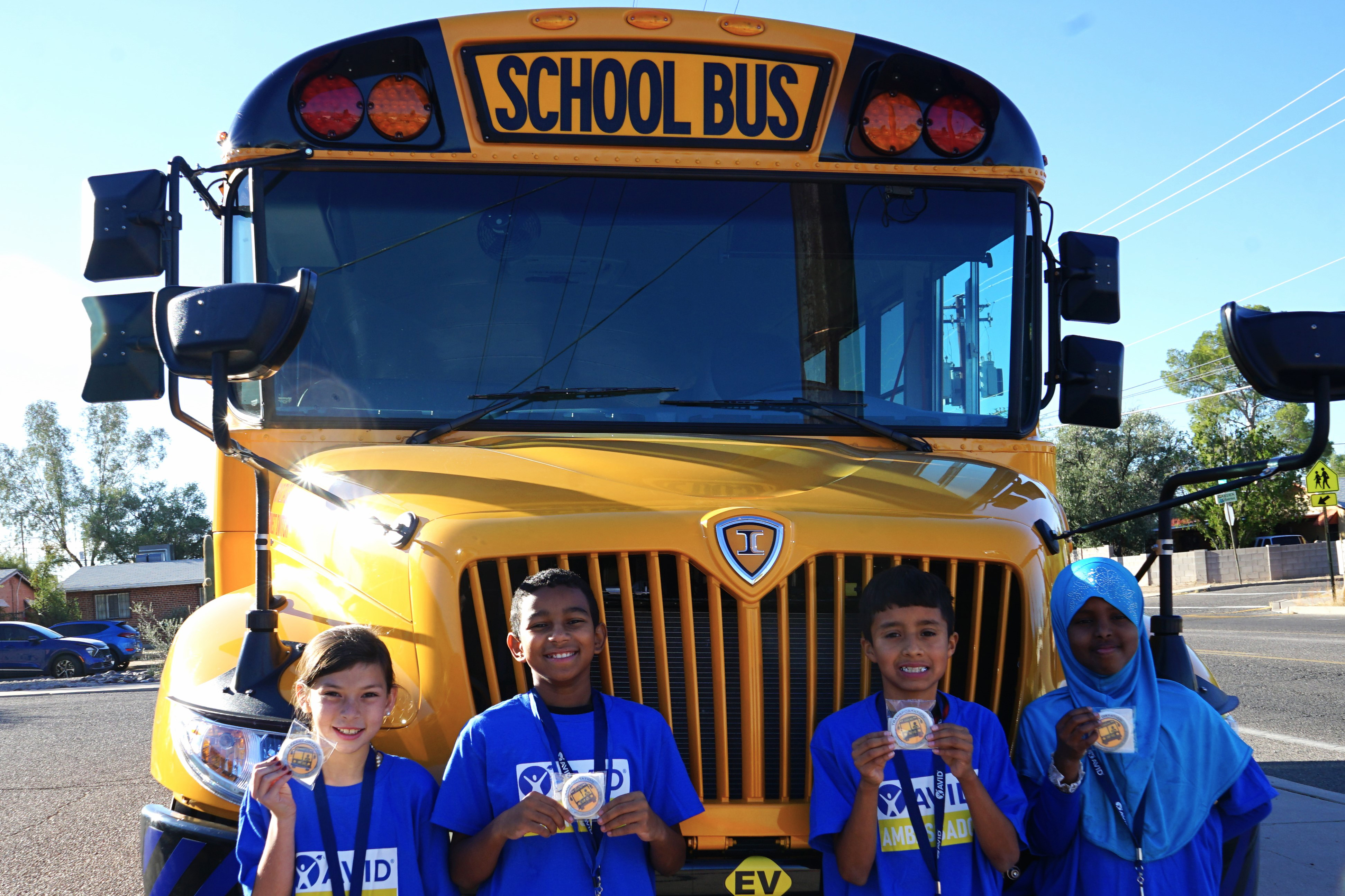 Four students in blue shirts smile in front of a new EV school bus