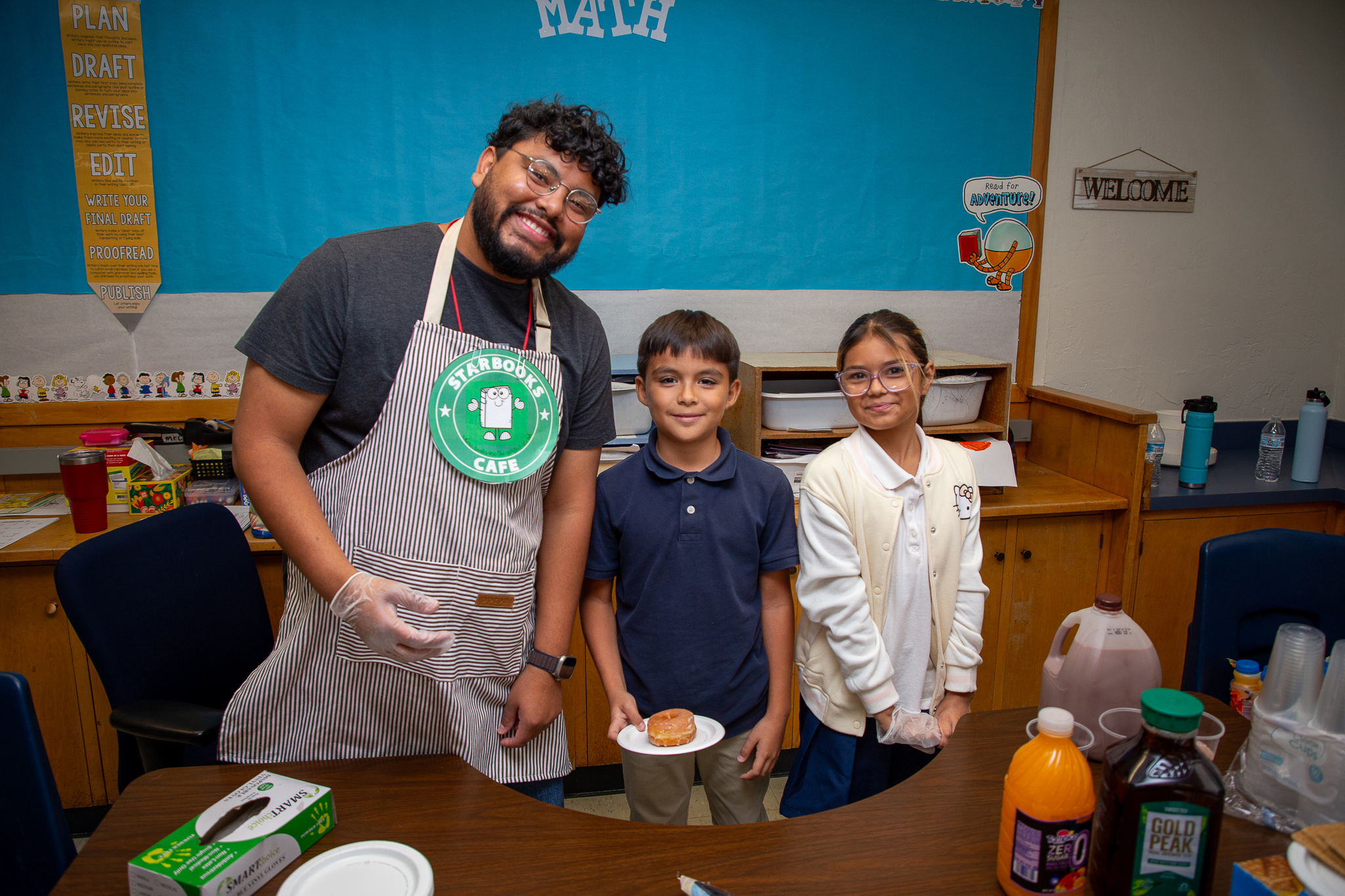 A teacher in an apron and two students smile behind a table filled with treats and drinks