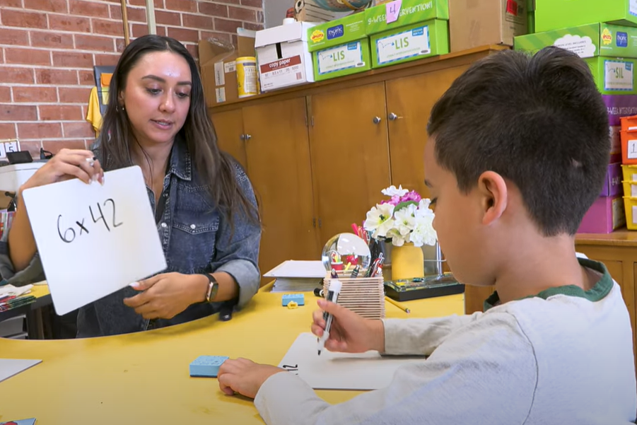 A teacher holds up a white board with 6 x 42 written on it to show a young boy