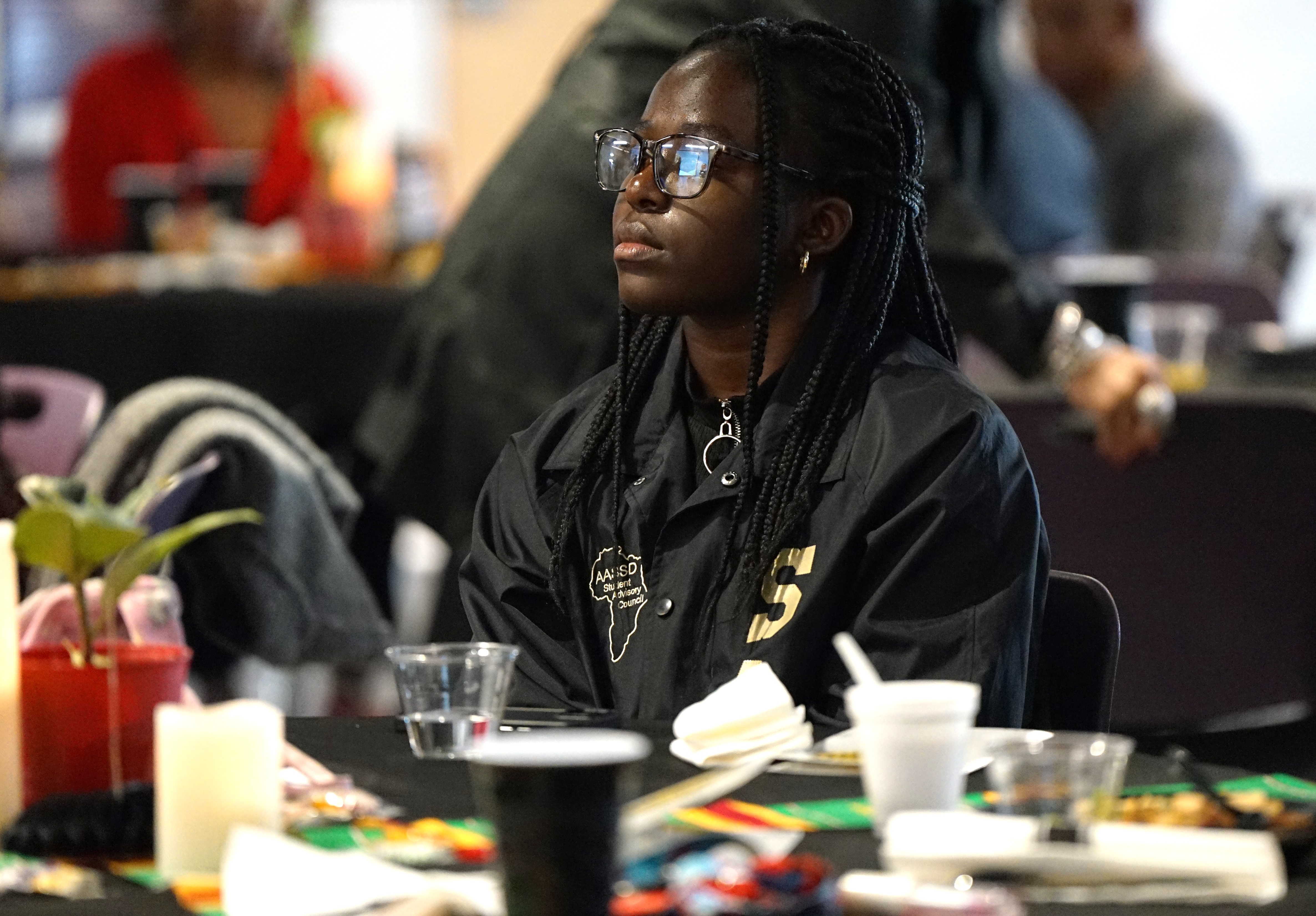 A girl in glasses sits at a table watching the presentation