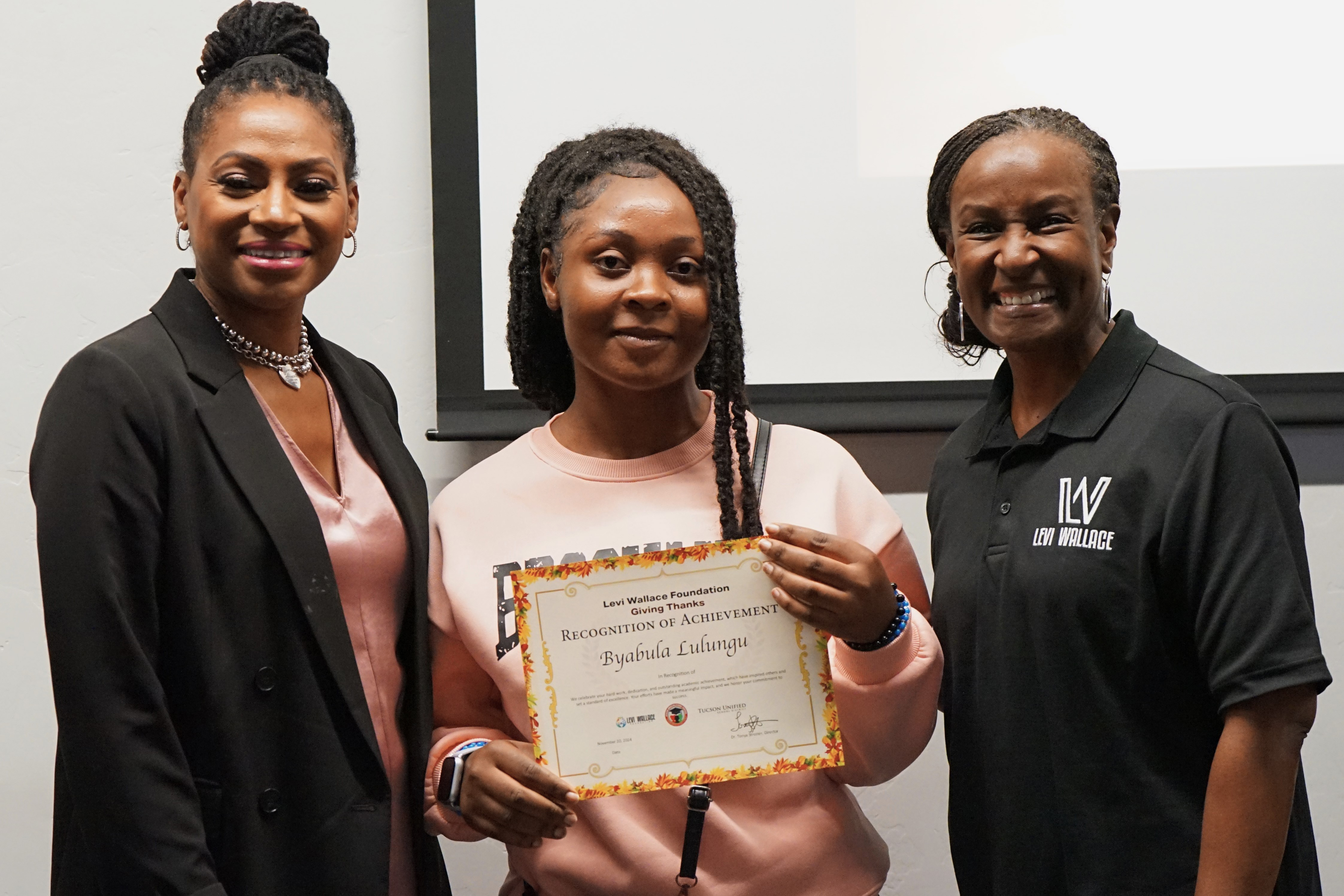 A young woman in pink holds a certificate in between two women in black