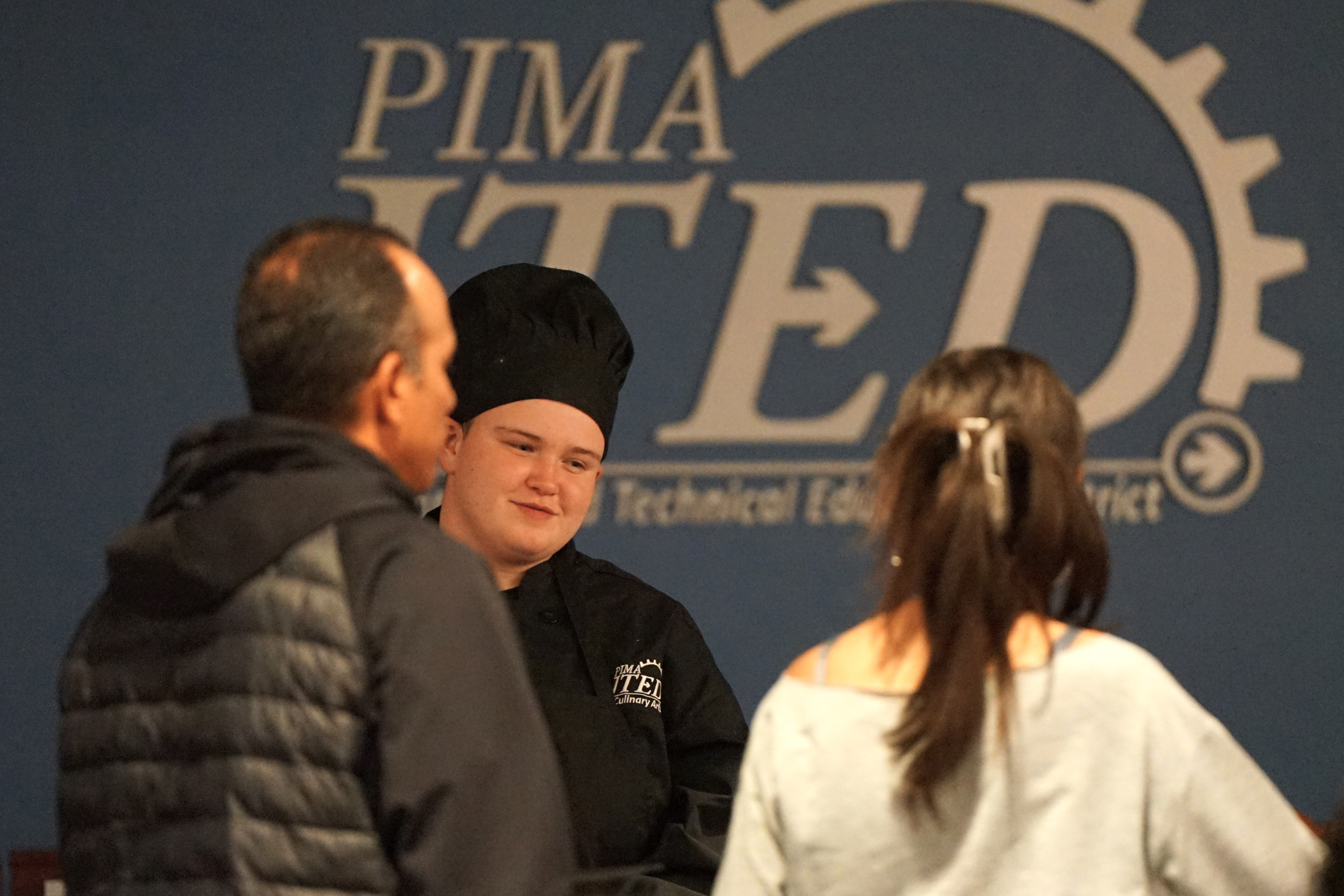 A student in a black chef hat serves food to a man and woman