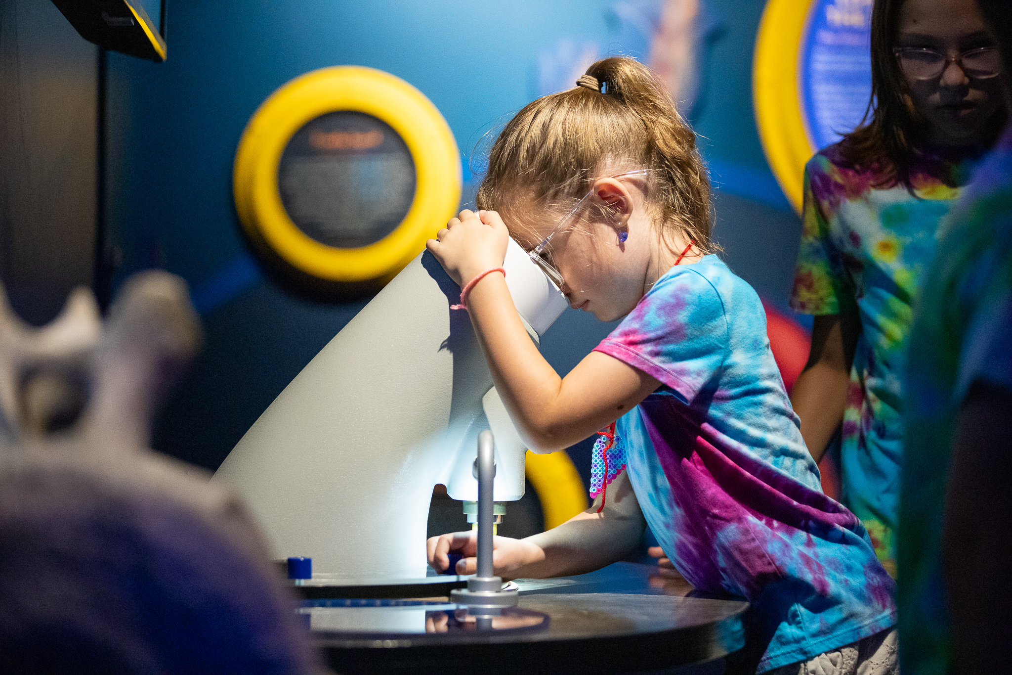 A little girl with glasses and a tie-dye shirt looks through a microscope