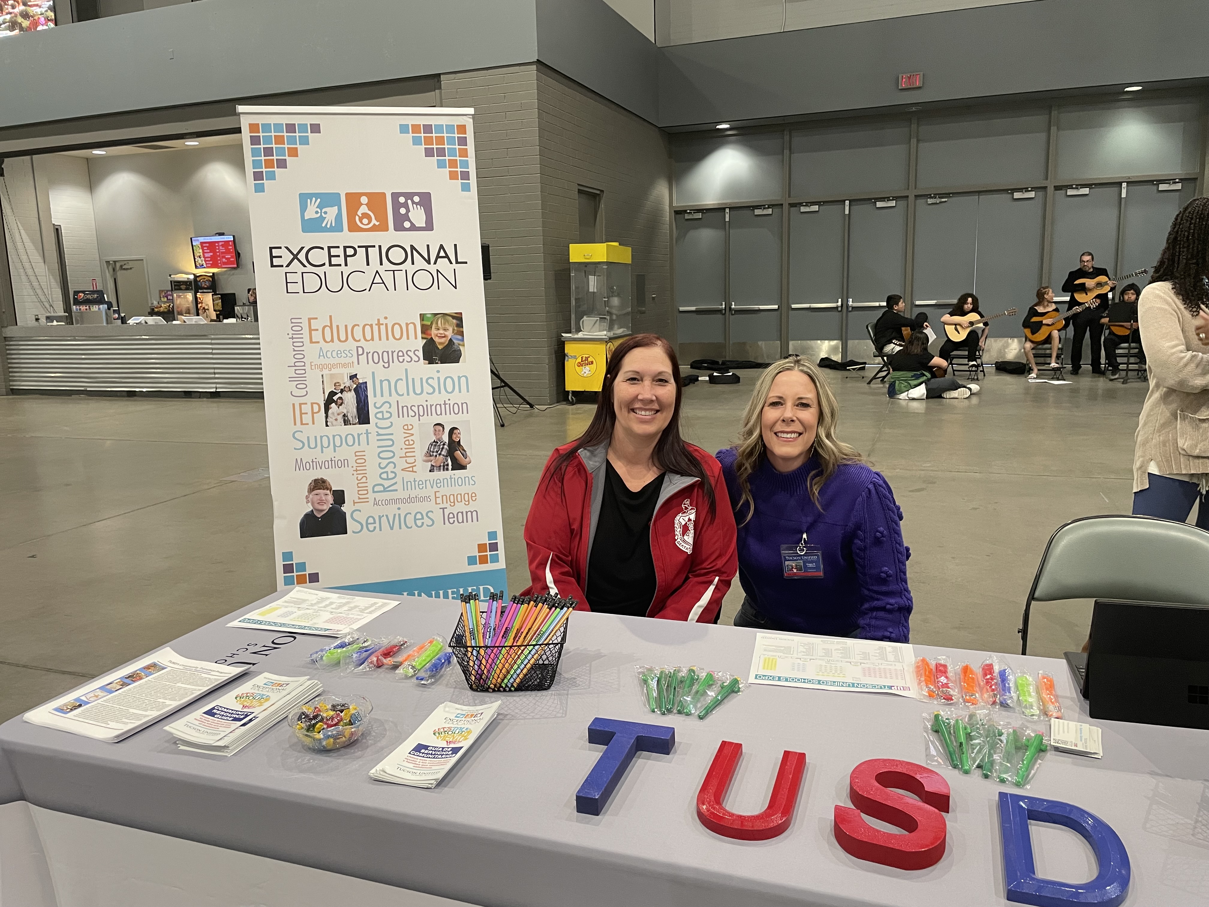 Two women smile at a table with TUSD letters on it