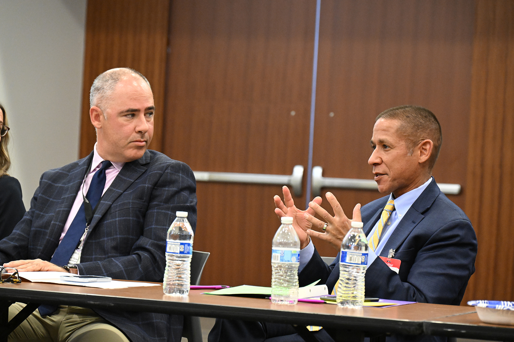 A man in a navy suit talks while a man in a black checkered suit listens