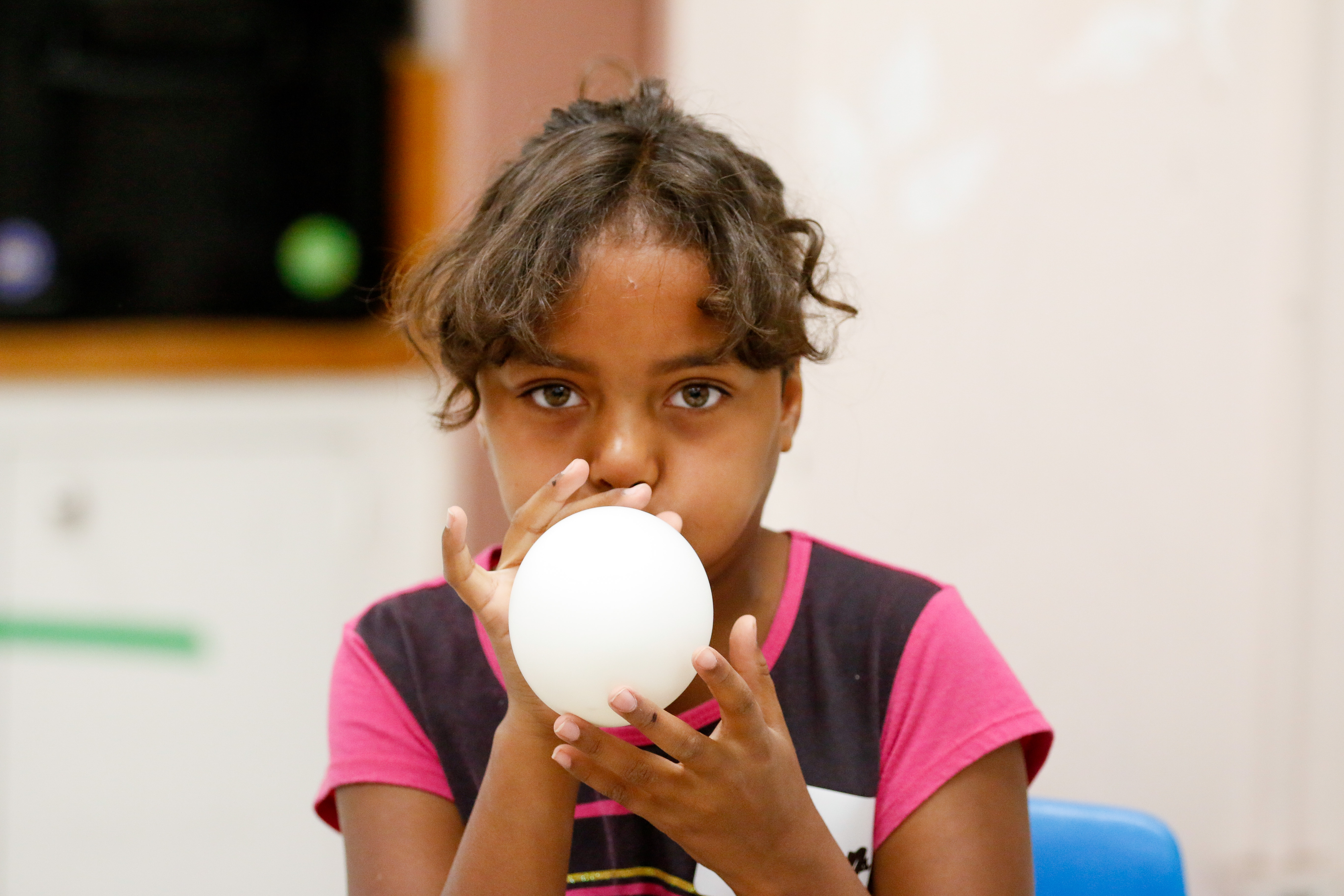 A little girl with dark brown curly hair blows up a white balloon