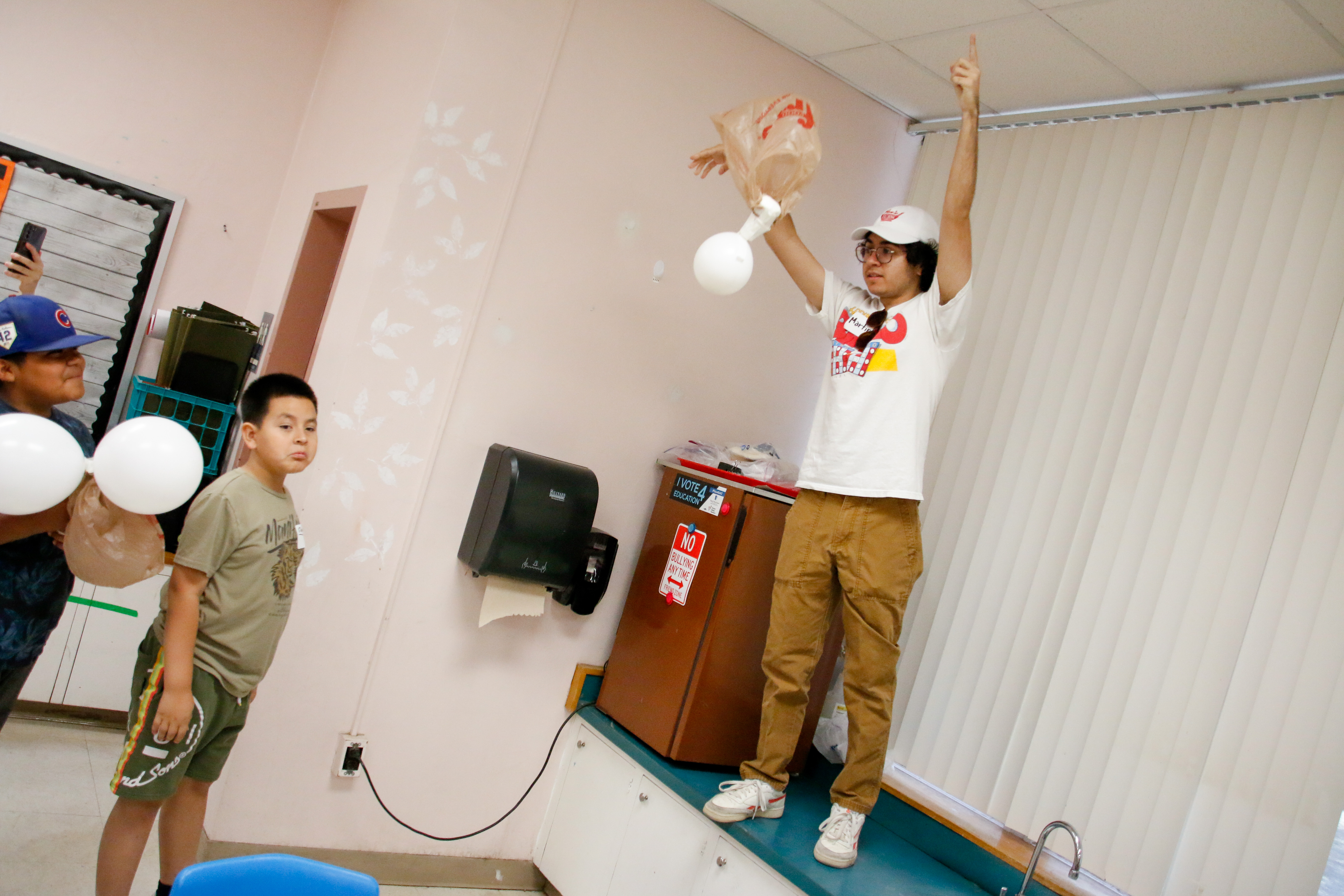 A boy in a white hat and shirt stands on a counter to drop his egg with a balloon and grocery bag