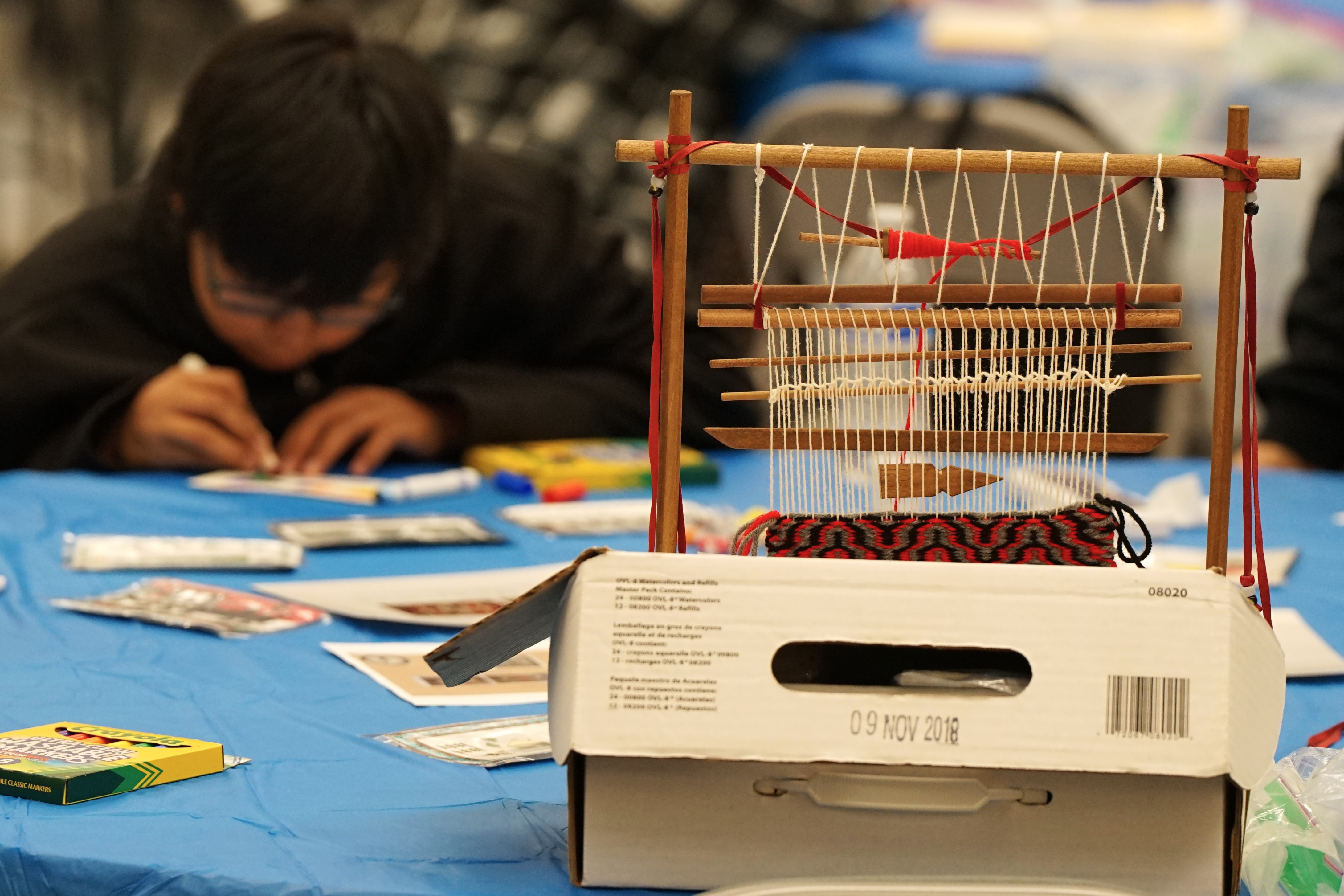 A boy wearing glasses colors with a marker, with a Native yarn craft in the foreground