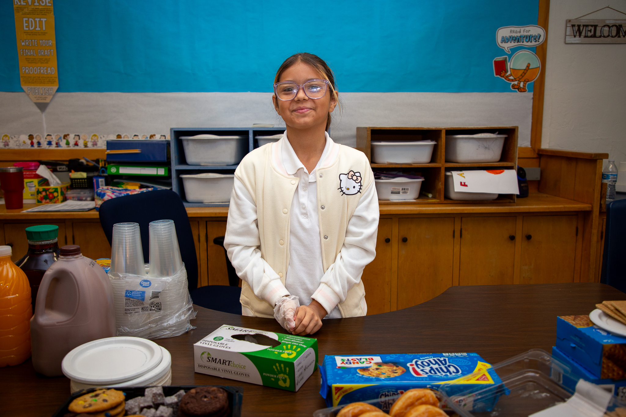 A girl in glasses and a white Hello Kitty jacket smiles standing behind a table with beverages and snacks