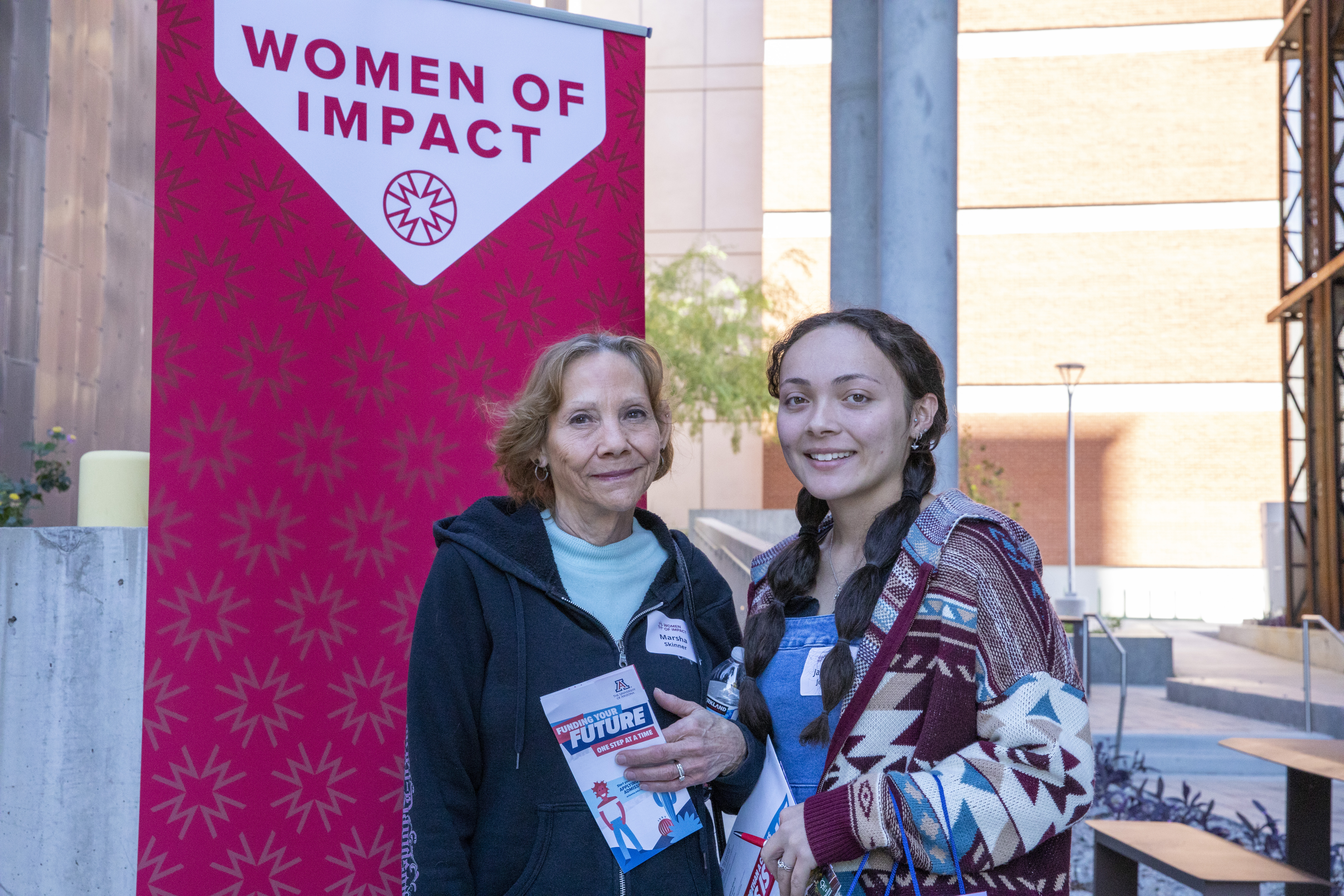 A teen girl smiles with her mentor in front of a red Women of Impact banner