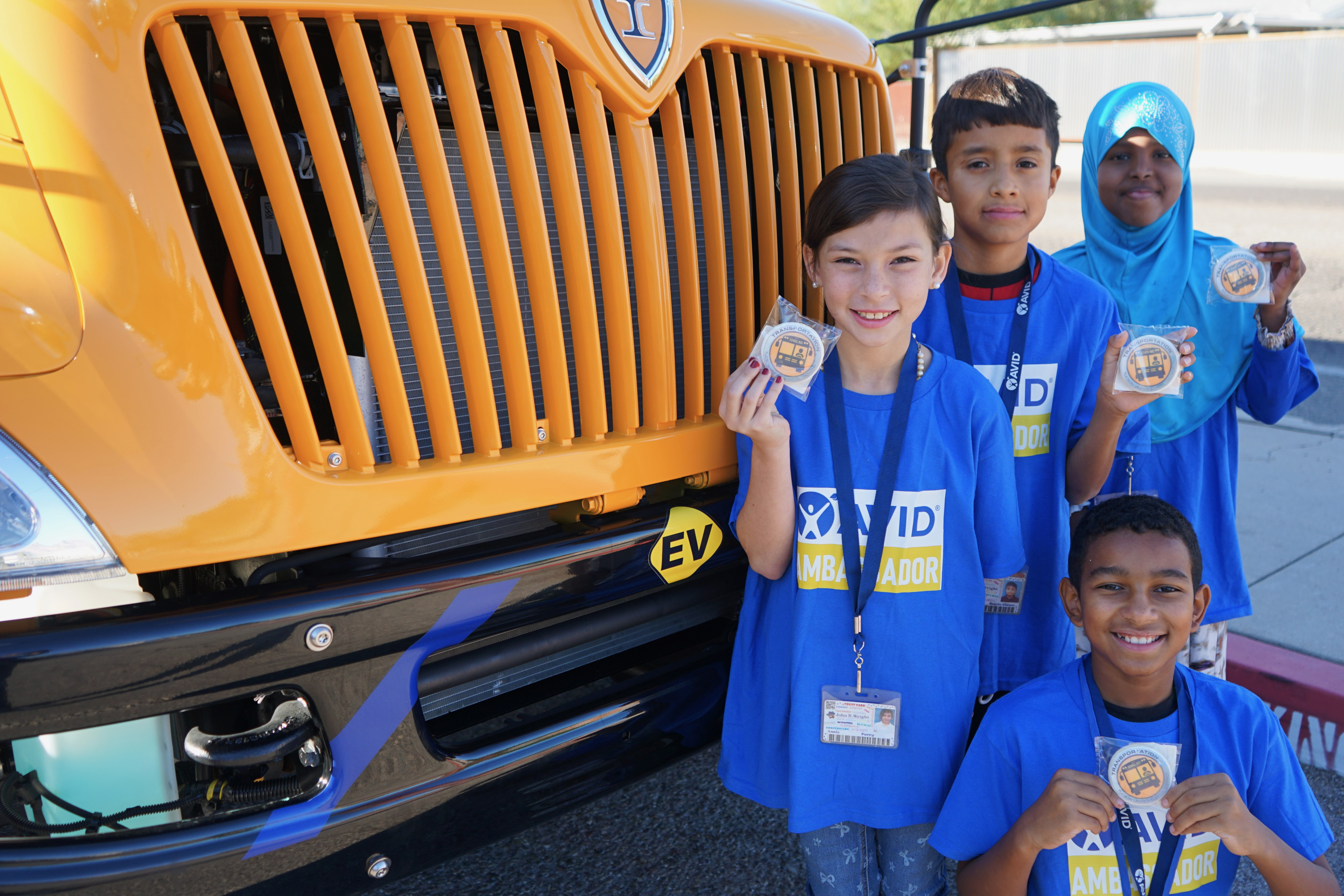 Four students in blue AVID shirts hold up cookies in front of one of the new EV school buses